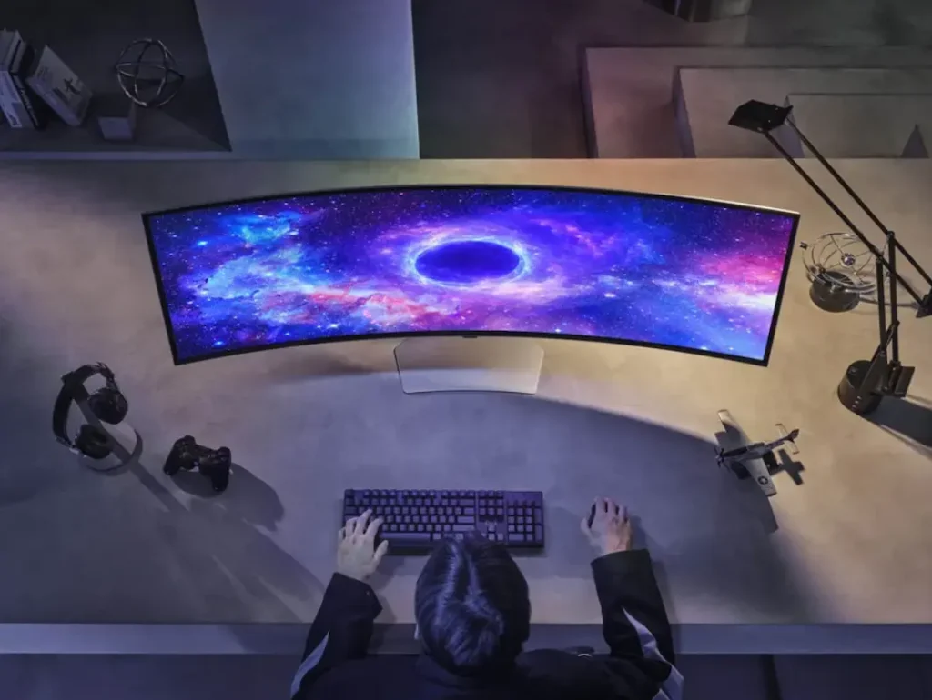 Visão frontal de um monitor curvo widescreen em um setup gamer escuro, com iluminação de fundo em LED e acessórios tecnológicos sobre a mesa.