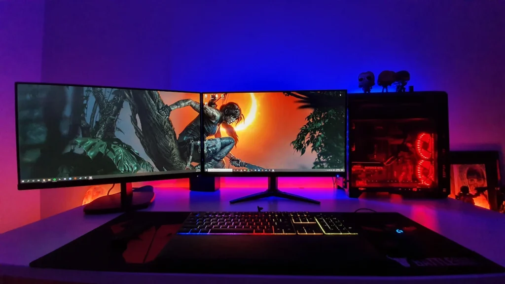 Setup gamer com dois monitores exibindo jogo de aventura, teclado com iluminação RGB e gabinete de computador com luzes vermelhas em ambiente escuro com iluminação azul e vermelha.